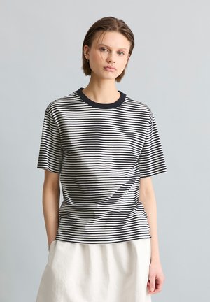 Young person with short hair wearing a black-and-white striped t-shirt and a white skirt, standing against a plain light gray background.