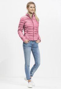 Lichtroze, puffy, trainingsjack met rits en hoge kraag en horizontale lijnen. Gepaard met lichtblauwe skinny jeans en witte sneakers.
