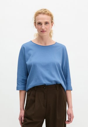 Kobieta o kręconych blond włosach, ubrana w niebieską bluzkę z rękawami do łokcia i wysokimi brązowymi spodniami, stojąca na tle białego tła.