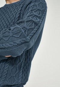 Pull en maille bleu marine avec motifs texturés et détails en torsades sur les manches. Col rond et manches longues.