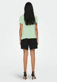 Hellgrünes Kurzarmoberteil mit lässiger Passform, kombiniert mit schwarzen Shorts und schwarzen High Heels, Ansichten von hinten.