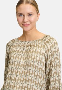 Blusa a maniche lunghe color marrone chiaro con un motivo astratto verde, rosa e bianco. Presenta un collo arricciato e una texture di tessuto morbido.