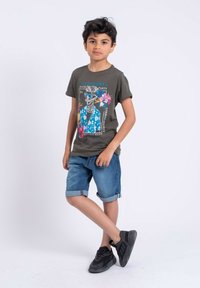 T-shirt vert foncé avec un graphique de squelette coloré, associé à un short en denim retroussé. Le garçon porte des chaussures de sport noires.