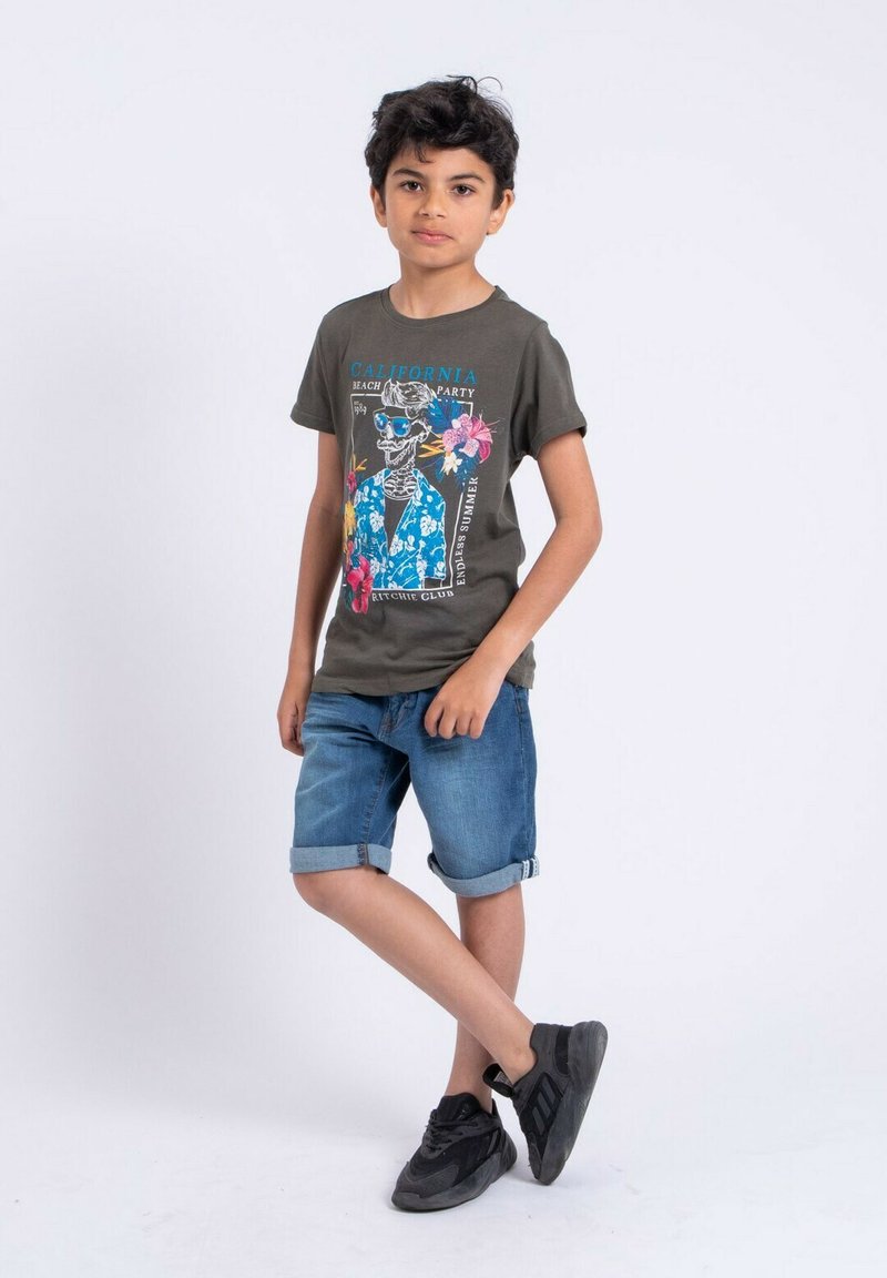 T-shirt vert foncé avec un graphique de squelette coloré, associé à un short en denim retroussé. Le garçon porte des chaussures de sport noires.