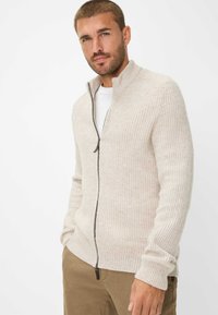 Beige geripptes Strickcardigan mit hohem Kragen, vollem Reißverschluss und strukturierter Oberfläche. Getragen über einem weißen T-Shirt und kombiniert mit khakifarbenen Hosen.