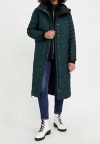 Abrigo largo acolchado en verde oscuro con capucha, con cremallera frontal y botones de presión. Lleva puesto sobre una chaqueta negra con cremallera y vaqueros, con botas blancas.