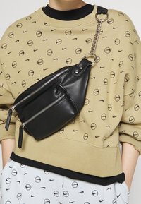 Svart faux läder crossbody-väska med två dragkedjefickor, ett blänkande kedjestropp och en stilren, vinklad design. Accenter inkluderar dragkedjehantag.