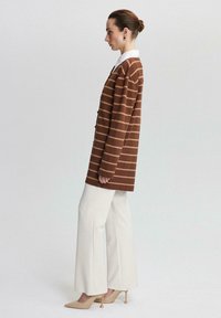 Bruine gestreepte lange cardigan met een witte kraag, gecombineerd met brede crèmekleurige broek en puntige beige hakken, tegen een neutrale achtergrond.
