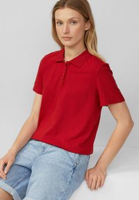 Polo rouge avec un col classique, manches courtes et une patte de boutonnage. Associé à un short en jean bleu clair, sur un fond neutre.