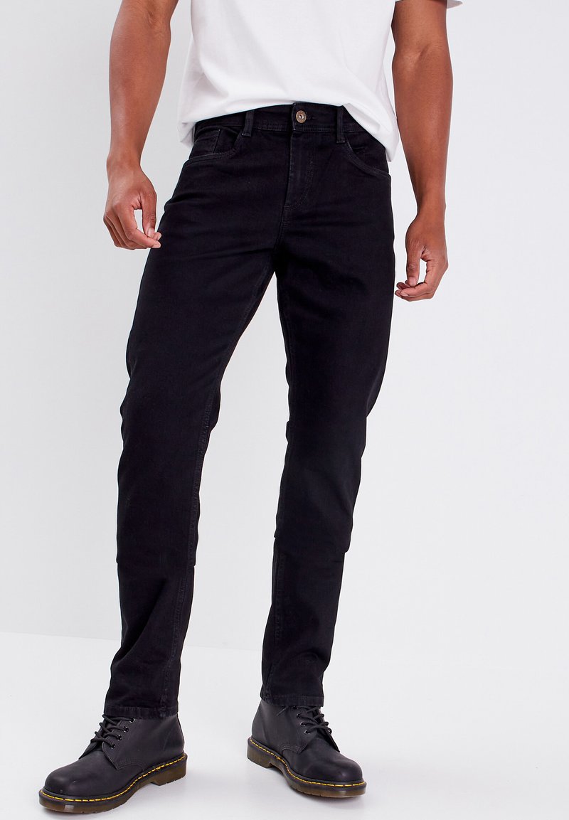 Schwarze Jeans aus Denim mit schmalem Schnitt, Knopfverschluss, Fronttaschen und dezenten Nähdetails. Tragen mit schwarzen Stiefeln.