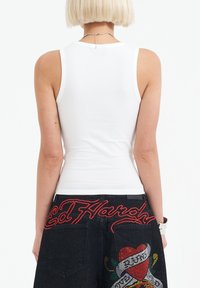 Witte aansluitende tanktop met smalle bandjes, gecombineerd met zwarte spijkerbroeken met rode borduursels en versieringen op de achterzakken.