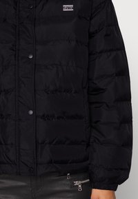 Giubbotto a sacco nero con texture trapuntata, bottoni a pressione e un piccolo logo. Presenta polsini elasticizzati e una tasca con zip sul lato.