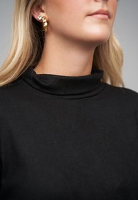 Zwarte coltrui met een zachte textuur en een omslagskraag. Gouden oorbellen in de vorm van ringen en bloemenaccenten versieren de halslijn.