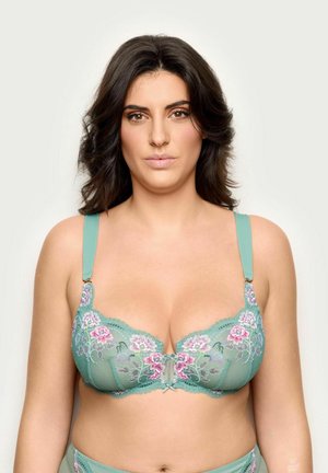 Donna con capelli scuri e mossi che indossa un reggiseno in pizzo turchese con ricamo floreale rosa, rivolta in avanti su uno sfondo chiaro uniforme.