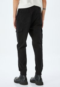 Schwarze Cargo-Hose mit elastischen Bündchen, ausgestattet mit Seitentaschen und hinteren Paspeltaschen. Hergestellt aus strapazierfähigem Material mit lockerem Schnitt.