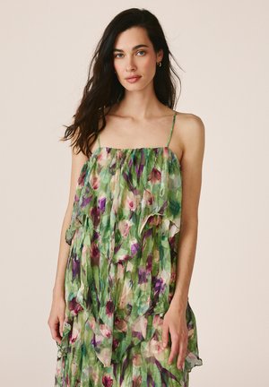 Mujer con cabello largo y oscuro que lleva un vestido verde con flores sin mangas y con tirantes finos sobre un fondo beige liso.