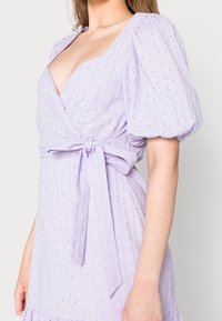Lavendelfärgad klänning med puffärmar, omslagsdesign och knytliv. Har broderade blommönster och en texturerad tyg.
