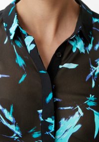 Blouse noire transparente ornée de motifs abstraits teal et bleus, avec une texture douce, un col classique et des détails de boutons sur le devant.