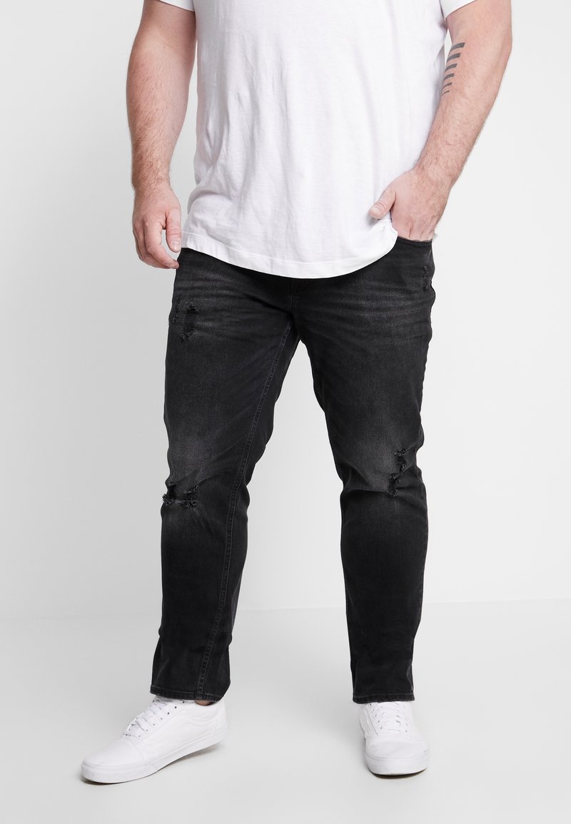 Vaqueros de mezclilla negra con detalles desgastados en la parte frontal, corte slim, combinados con una camiseta blanca y zapatillas blancas.