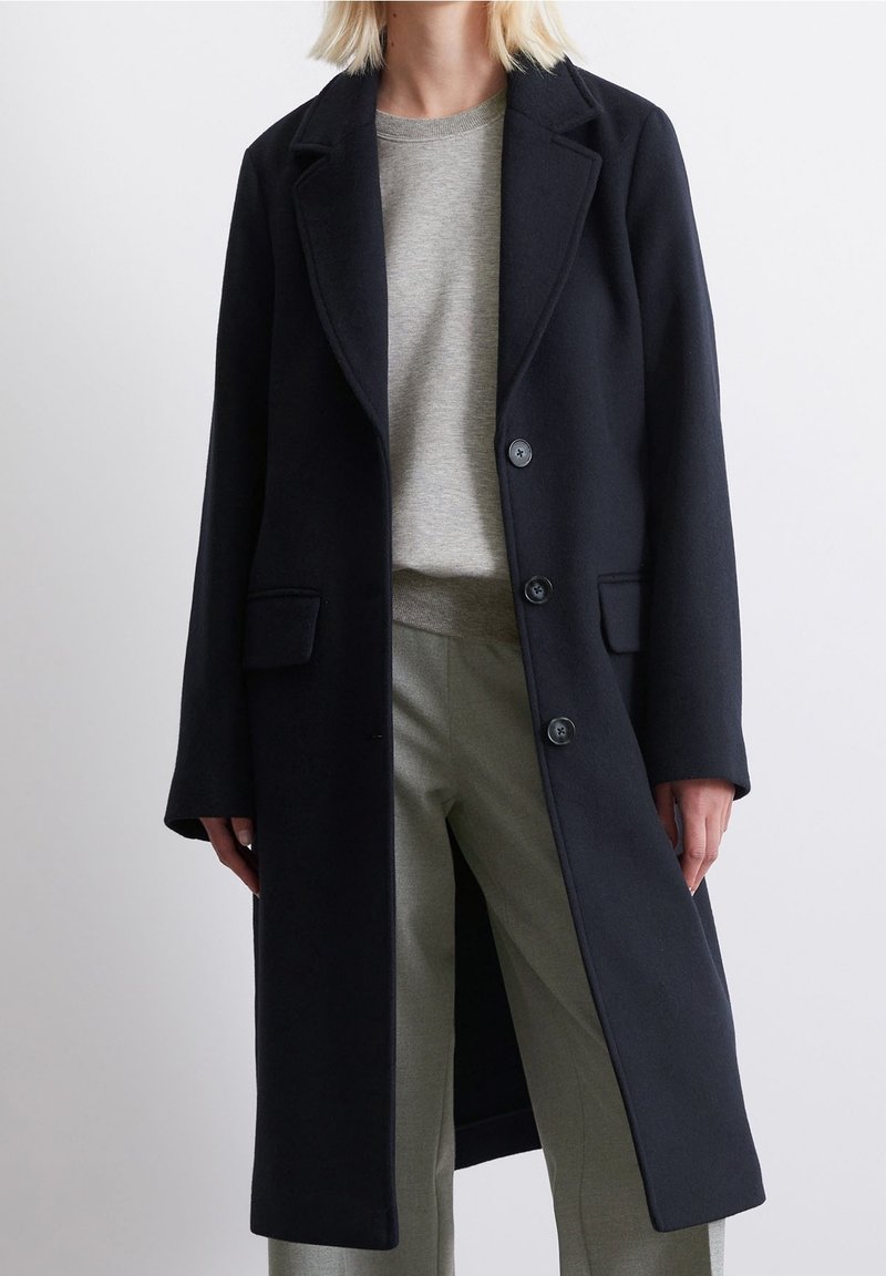 Abrigo de lana negro con solapas muesca, diseño de un solo botón y dos bolsillos frontales. Combinado con un suéter gris claro y pantalones verde claro.