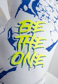 Tessuto bianco con motivi grafici blu presenta il testo "ESSERE L'UNICO" in un brillante neon giallo, testurizzato e in grassetto.