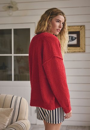 Sieviete ar viļņainiem blondiem matiem, valkājot teksturētu sarkanu džemperi un melnbaltas svītrainas šortus gaišā, modernā istabā.