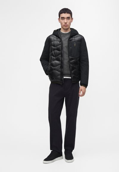 Jeune homme portant une veste matelassée noire à capuche, un pull gris, un pantalon noir et des baskets noires à semelles blanches, debout sur un fond blanc.
