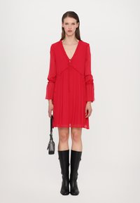 Robe en chiffon rouge avec un décolleté en V, des détails en volants et des manches longues. Associée à des bottes en cuir noires jusqu'aux genoux et à un petit sac à main noir.