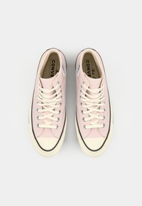 Lyse rosa lerret sneaker med kremfargede snørebånd og off-white tåhette, med to sirkulære logo-patcher på sidene og svart gummisåle.