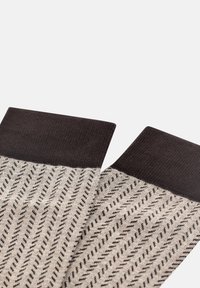 Adītas zeķes ar melnu ribotu aproci, krēmkrāsas pamatu ar abstraktiem melniem diagonāliem svītrojumiem, uzsverot tekstūras un raksta kontrastu.