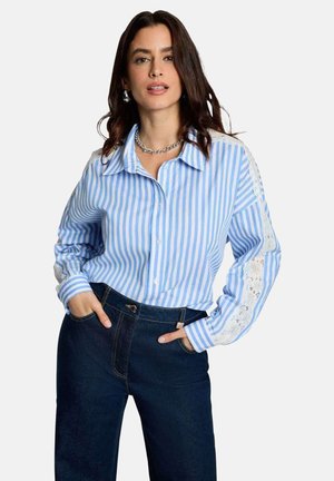 Femme portant une chemise à rayures verticales bleues et blanches avec des détails en dentelle sur les manches et un jean taille haute bleu foncé, les mains dans les poches.