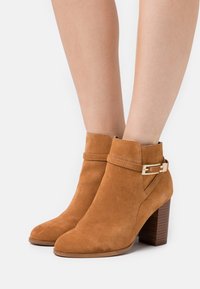 Botas de tobillo de ante en color marrón claro con un acabado texturizado, que presentan un detalle de hebilla y un tacón de madera apilada. Diseño de punta redondeada.