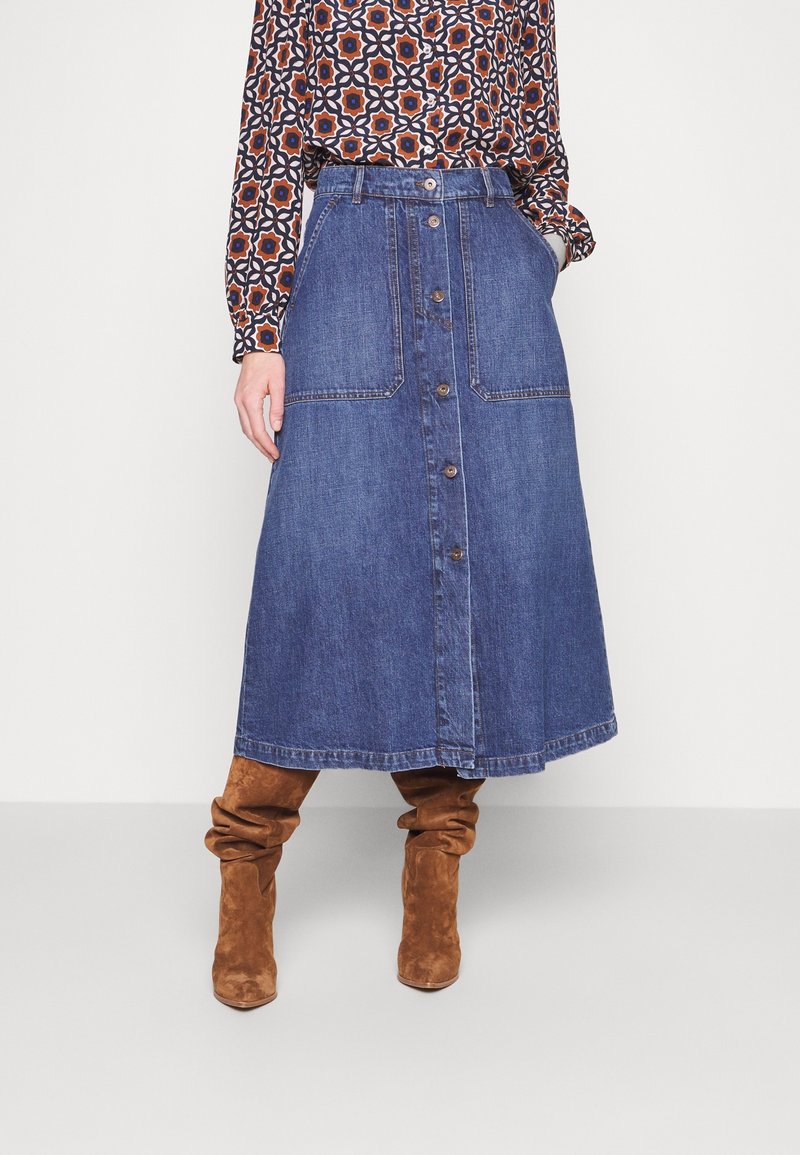 Femme portant une jupe midi en jean bleu à boutons sur le devant, une chemise à manches longues à motifs et des bottes en daim marron souples.