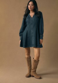 Tumša džinsa kleita ar V veida izgriezumu, gariem pūkainiem piedurknēm un sānu kabatām; kombinēta ar ceļos sasniedzamām brūnganām zamšādas zābakām.