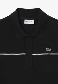 Musta polo-paita, joka on valmistettu teksturoidusta kankaasta. Siinä on lyhyt kaulus, kolme nappia, kontrastivalkoiset raidat ja vihreä krokotiililogo.