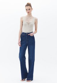 Béžový bodysuit s detailním zipem, spárovaný s tmavě modrými širokými džíny s vysokým pasem, které mají minimální švy a klasický pětikapsový design.