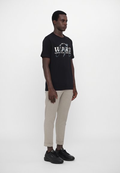 Tricou din bumbac negru cu grafică "WRONG PLACE RIGHT TIME" în alb și albastru, asortat cu pantaloni tapered bej și adidași negri.