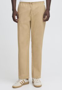 Beige cotton pants with a relaxed fit, featuring an elastic waistband and drawstring. Paired with white sneakers and subtle striped accents.