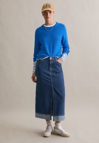 Niebieski sweter z długim rękawem z białymi i niebieskimi paskowanymi mankietami, połączony z długą spódnicą maxi z wysokim stanem z denimu i białymi trampkami. Beżowa czapka z daszkiem.