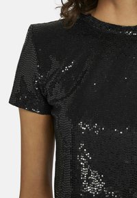 Zwarte sequined top met een gestructureerde afwerking, korte mouwen en een ronde hals. Glanzende sequins bedekken de stof en reflecteren het licht op verschillende manieren.
