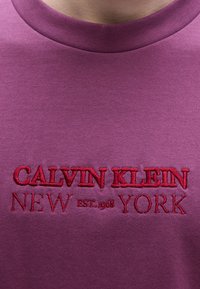 Μωβ βαμβακερό t-shirt με στρογγυλή λαιμόκοψη, με κόκκινο κεντημένο κείμενο στην μ προστινή πλευρά που γράφει "CALVIN KLEIN NEW YORK EST. 1968".