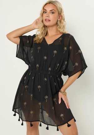 Donna che indossa un abito nero trasparente con stampe di palme dorate, maniche corte, vita aderente e nappe sull'orlo, posando con una mano sul fianco.