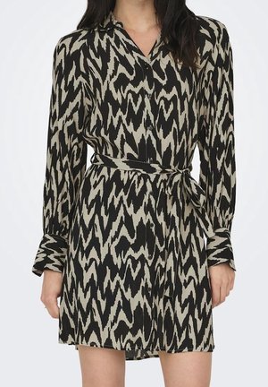 Femme portant une robe chemise à manches longues à motifs zigzag noirs et beiges, avec une ceinture en tissu nouée à la taille.