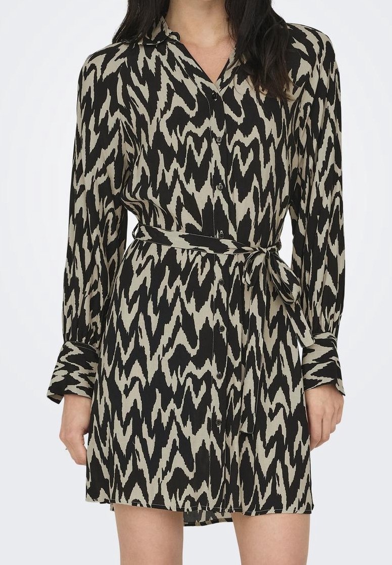 Femme portant une robe chemise à manches longues à motifs zigzag noirs et beiges, avec une ceinture en tissu nouée à la taille.