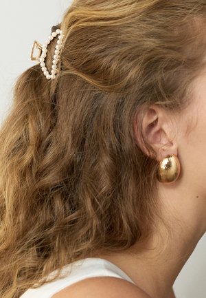 Curly light brown hair clipped back with a gold and pearl hair claw, shown with a large oval gold earring on the right ear.