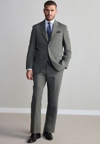 Mand iført grå jakkesæt med mørkeblå slips og brune sko, stående indendørs mod en ensfarvet lys grå væg og tæppebelagt gulv.