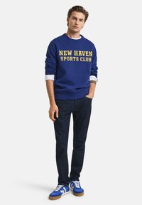 Blue sweatshirt with “NEW HAVEN SPORTS CLUB” in yellow text, white shirt cuffs visible, paired with dark jeans and blue sneakers with white accents.