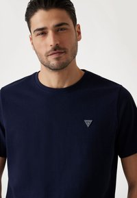 Marineblå bomulds t-shirt med rund hals og korte ærmer; har et trekantet logo-patch på venstre bryst. Glat tekstur.