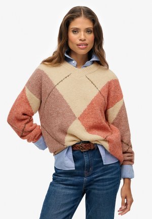 Maglione oversize con scollo a V in morbido tessuto, con un motivo a diamante nei colori crema, ruggine e pesca. Indossato sopra una camicia colletto azzurro chiaro.