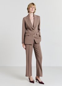 Abito marrone su misura con giacca monopetto, rever a U, tasche frontali e pantaloni dritti coordinati. Pumps borgogna con tacco alto.